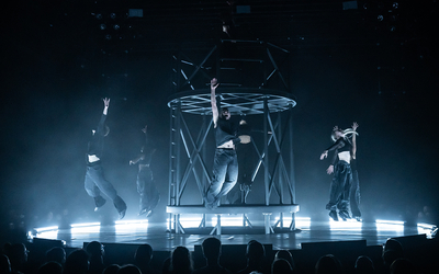 Une dizaine de danseurs tous habillés en noir, sautent sur une plateforme circulaire en métal éclairé par une lumière blanche. 