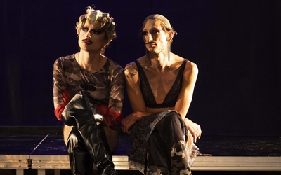 Deux artistes drags, l'un avec une permanente, sont assis les jambes croisées, regardant de manière pensive dans la même direction, les yeux dans le vague