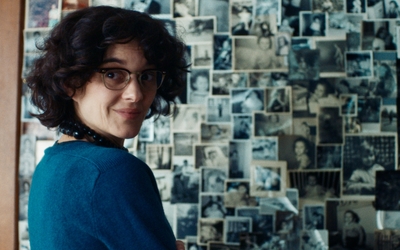 Une femme, la cinquantaine, se retourne vers nous, un sourire timide aux lèvres. Ele fait face à un mur de photos et de polaroïds