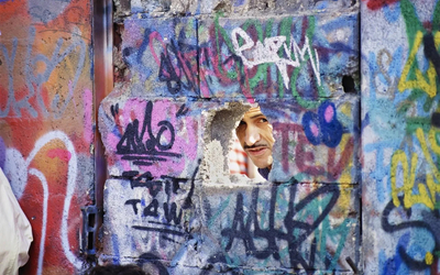 Mur de tags avec un trou d'où l'on voit apparaître le visage d'un homme moustachu