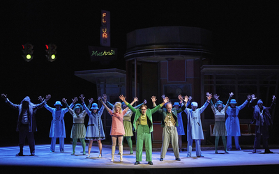 Sur une scène de théâtre, plusieurs personnes habillées de façon colorée lèvent les bras