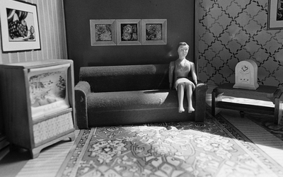 Photo en noir et blanc représentant une poupée de cire assise sur un canapé dans un salon des années 70
