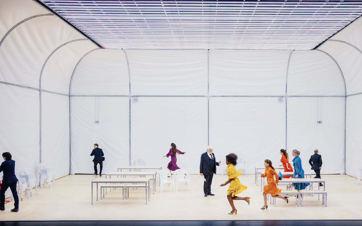 Photo de la pièce Les Conséquences. Jacques Weber est en costume bleu, des femmes en robes colorées courent de la droite vers la gauche