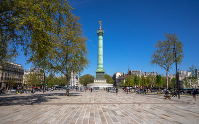 La Place de la Bastille à Paris, sous un ciel bleu dégagé. Au centre se dresse la Colonne de Juillet, un haut monument vert-de-gris surmonté de la statue dorée du Génie de la Liberté. La place est pavée de dalles claires et bordée d'arbres feuillus au printemps. Des bâtiments haussmanniens typiques entourent la place à l'arrière-plan. Quelques passants sont visibles marchant ou se reposant sur la place.