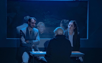 Décor de théâtre où l'on voit trois personnages : deux enquêteurs, et une femme blonde de dos qui semble interrogée