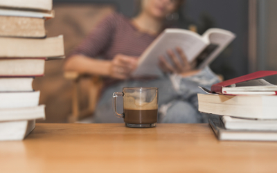 Table avec livres et tasse de café + une femme lit un ivre au loin (floue)