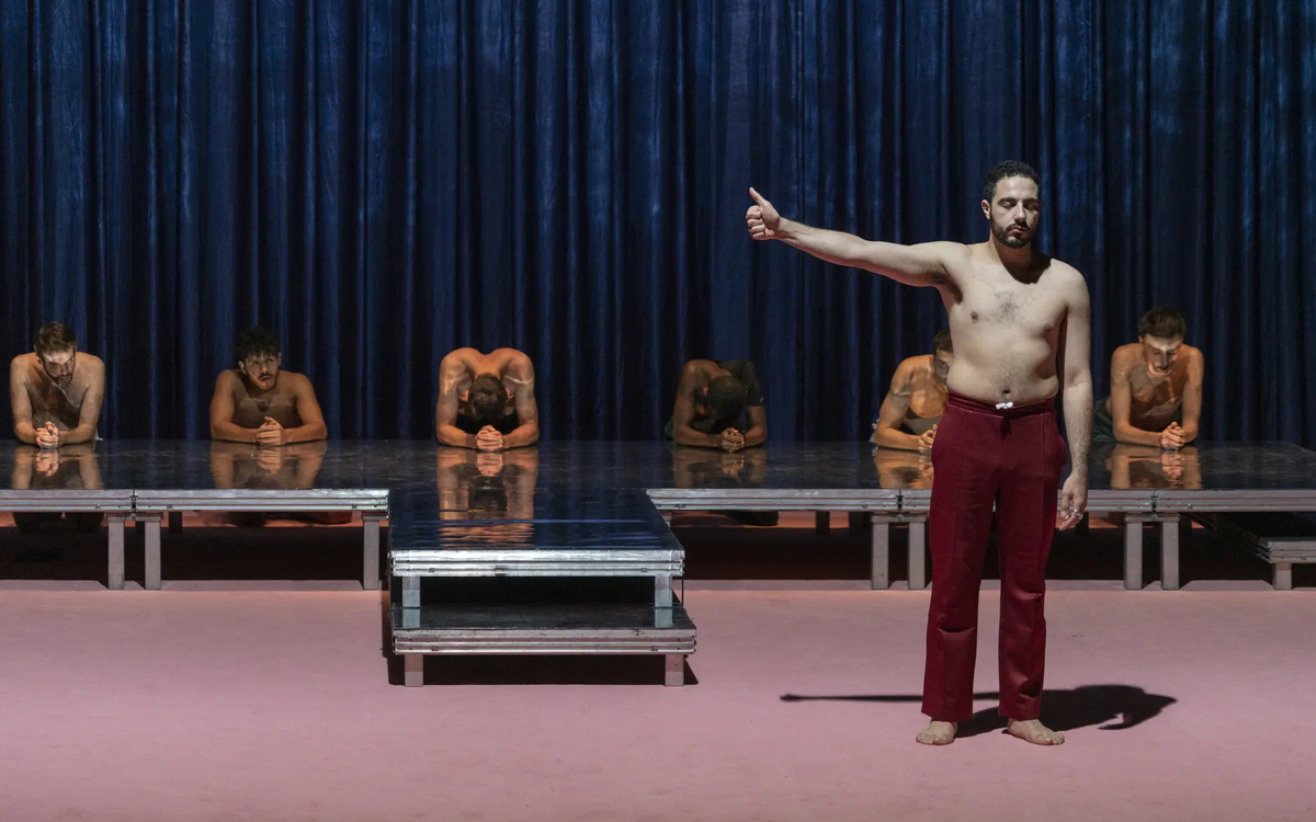 Sur une scène de théâtre, une rangée d'hommes accroupis, tête baissée et un homme à l'avant, torse-nu, pouce en l'air