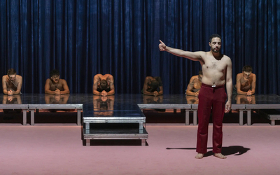 Sur une scène de théâtre, une rangée d'hommes accroupis, tête baissée et un homme à l'avant, torse-nu, pouce en l'air