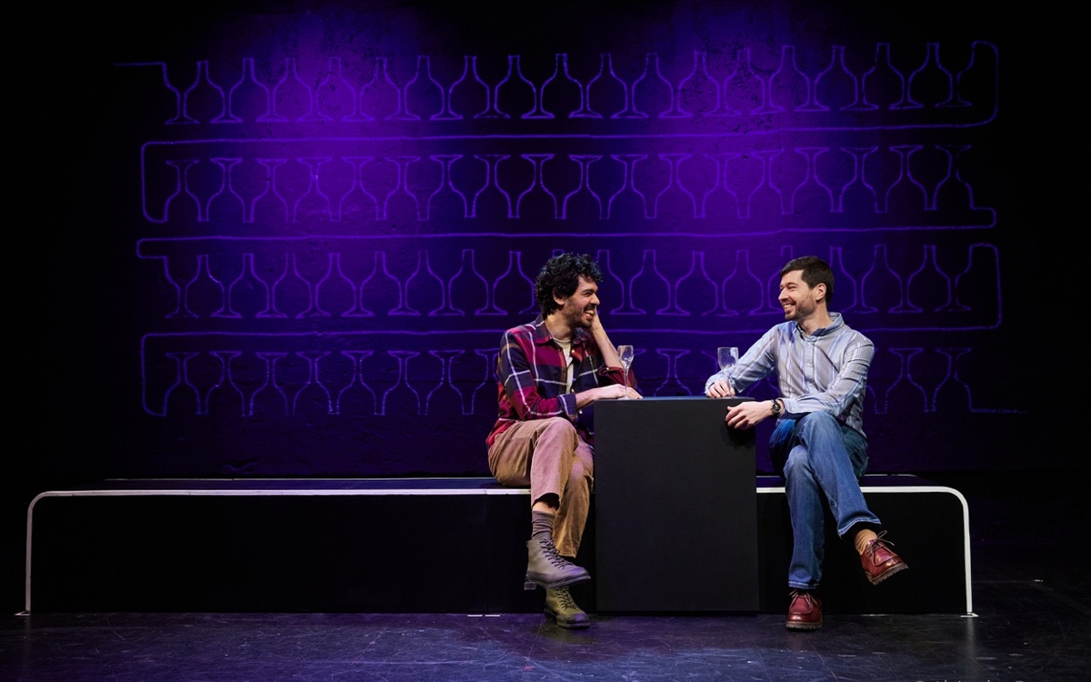 Sur une scène de théâtre, deux hommes boivent un verre en souriant