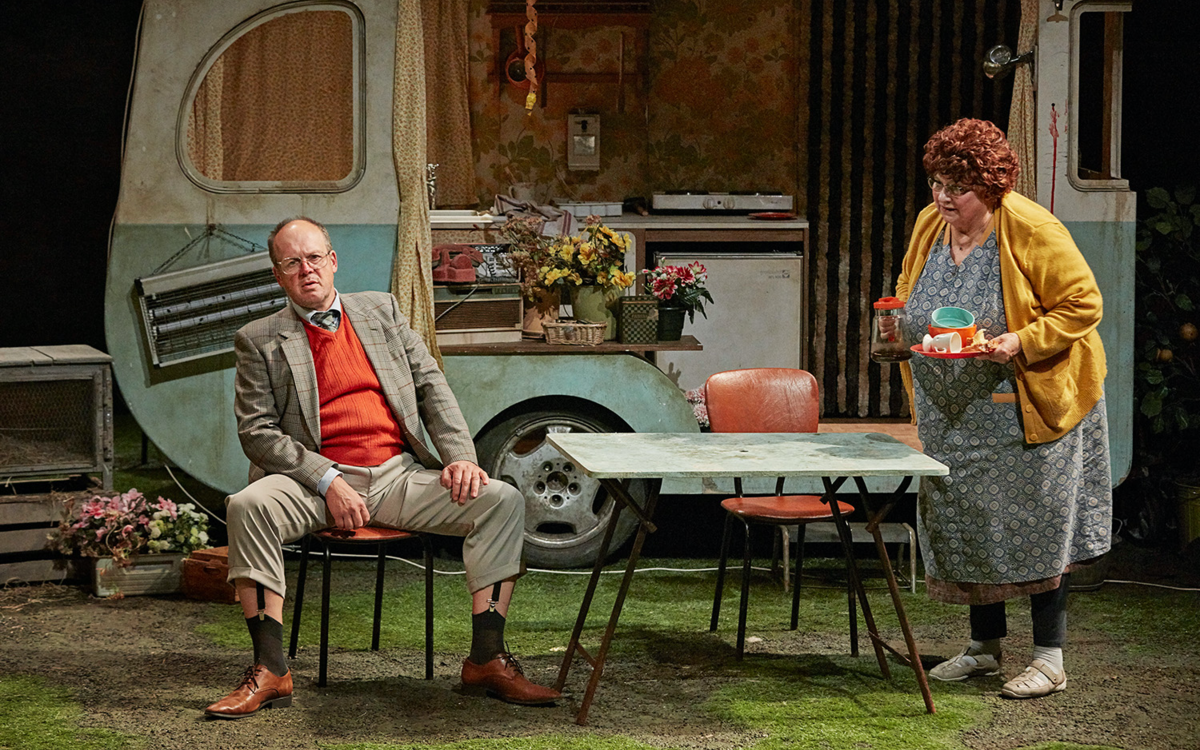 Sur une scène de théâtre, deux personnes âgés devant une roulotte vintage