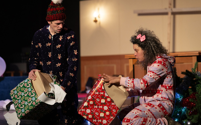 Sur une scène de théâtre, deux adultes glissés dans des pyjamas de noël ouvrent des cadeaux