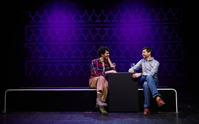 Sur une scène de théâtre, deux hommes boivent un verre en souriant