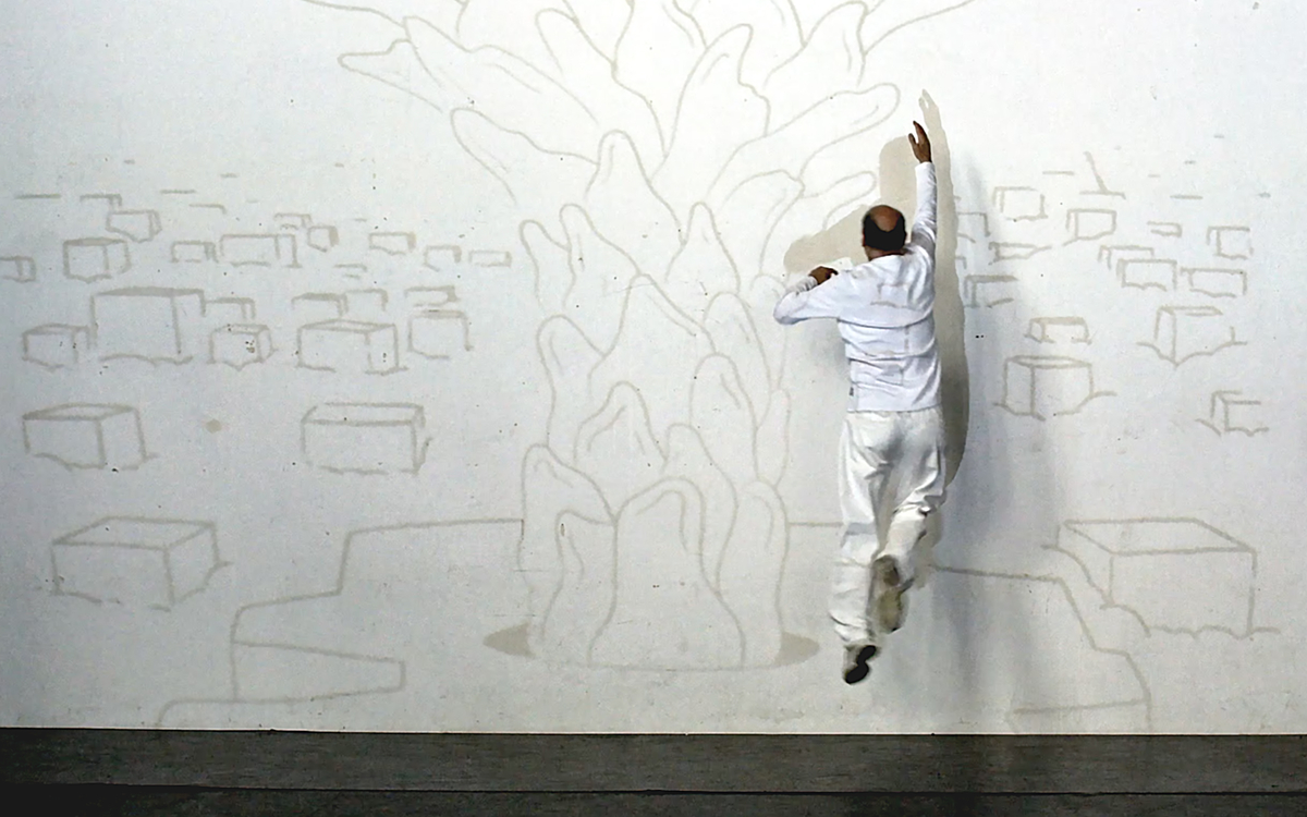 un homme habillé de blanc est face un mur où est dessiné un arbre et des rectangles pouvant ressembler à des maisons. 