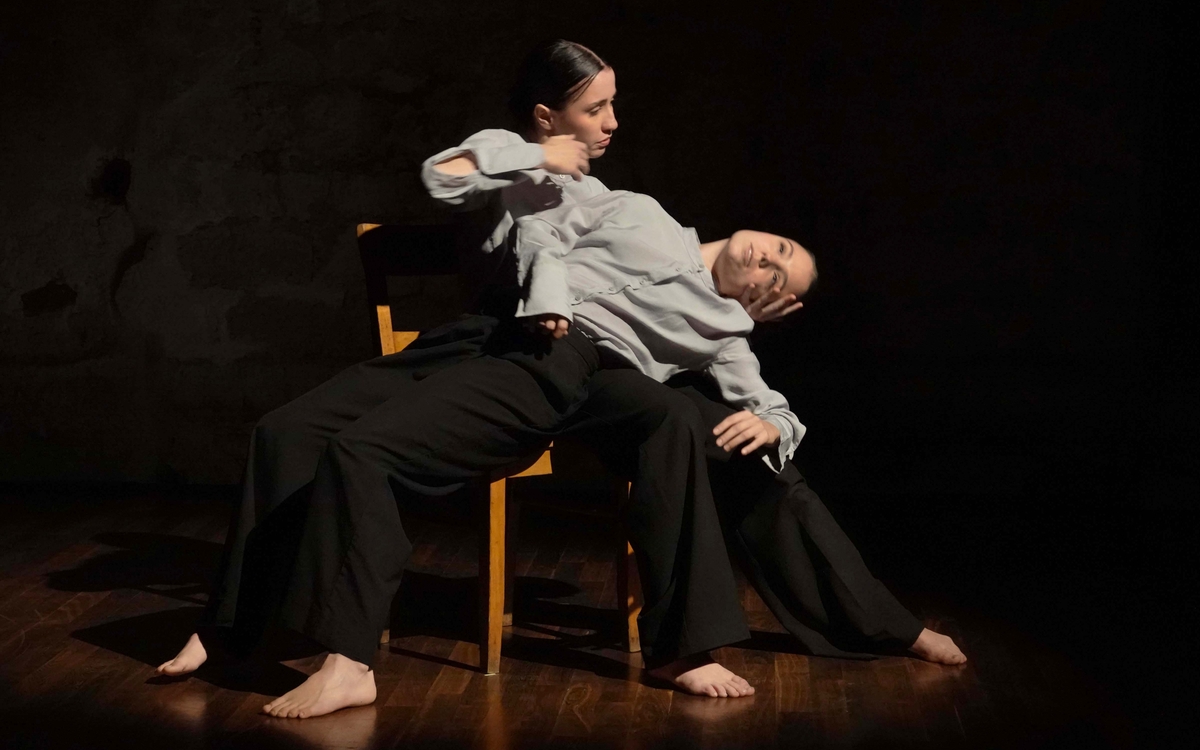 Deux femmes habillées d'une chemise grise et d'un pantalon noir sont sur une chaise. L'une est allongée sur les genoux de l'autre, qui lui tien le visage. 