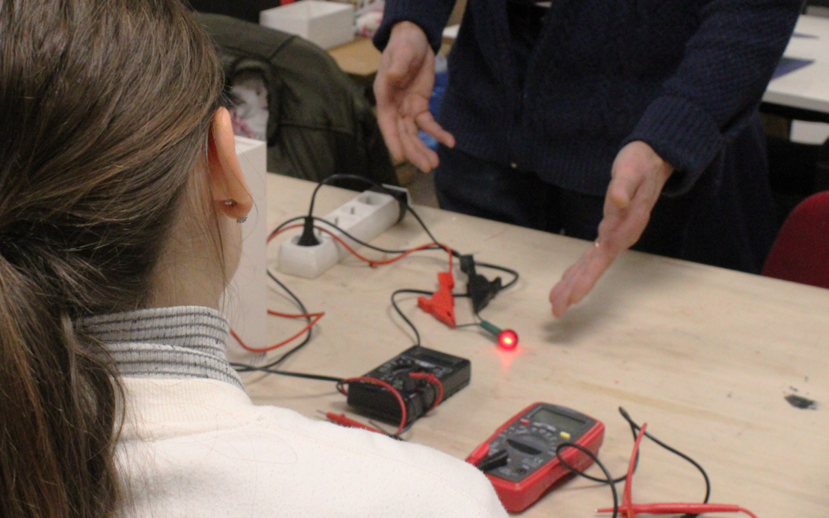 Photographie de dos d’une participante et des mains de l’intervenant lors d’un atelier d’initiation à l’électro.