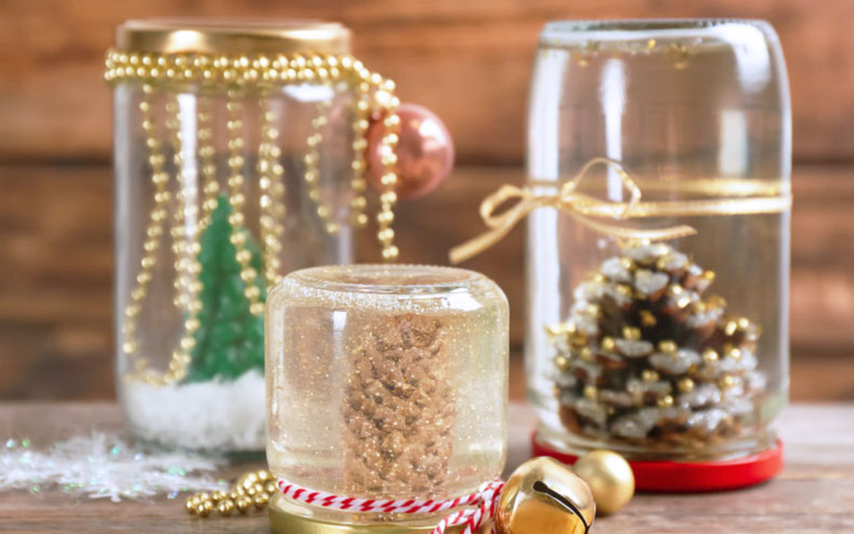 Photographie de 3 boules à neige sur le thème de Noël, faites dans des bocaux avec des pommes de pin, des paillettes et de l’eau.