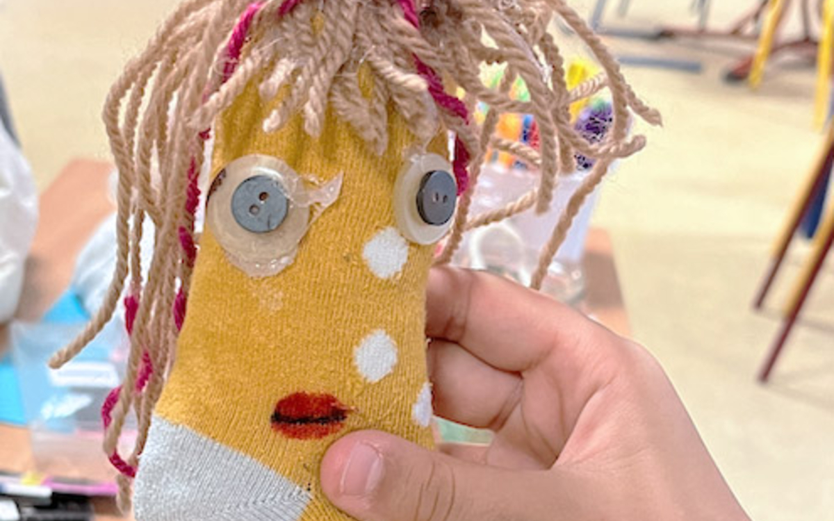 Photographie de la d’une petite fille tenant une peluche faite à partir d’une chaussette jaune à petits pois sur laquelle elle a collé des yeux en boutons et des cheveux rose et beige en laine.