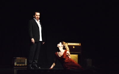 Sur une scène de théâtre, une femme dans une robe rouge aux pieds d'un homme habillé d'un costume