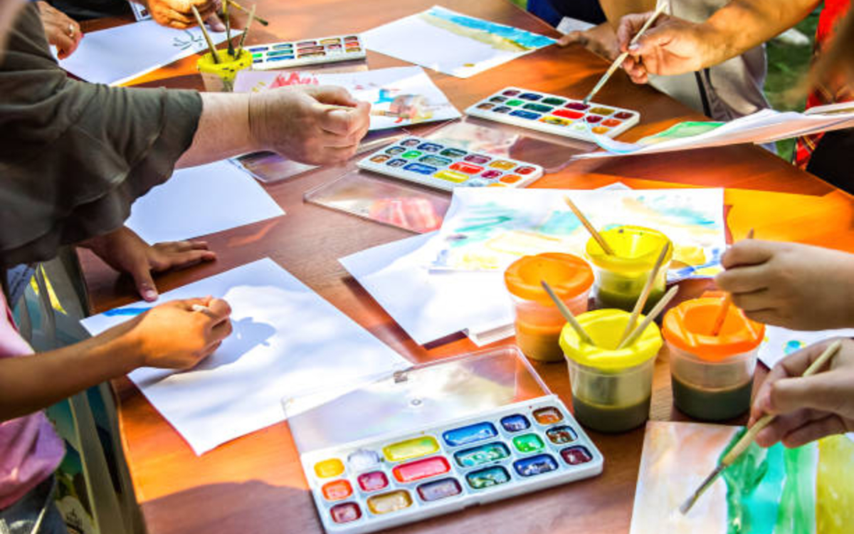 une table avec papier et palettes de peinture, des mains tiennent des pinceaux