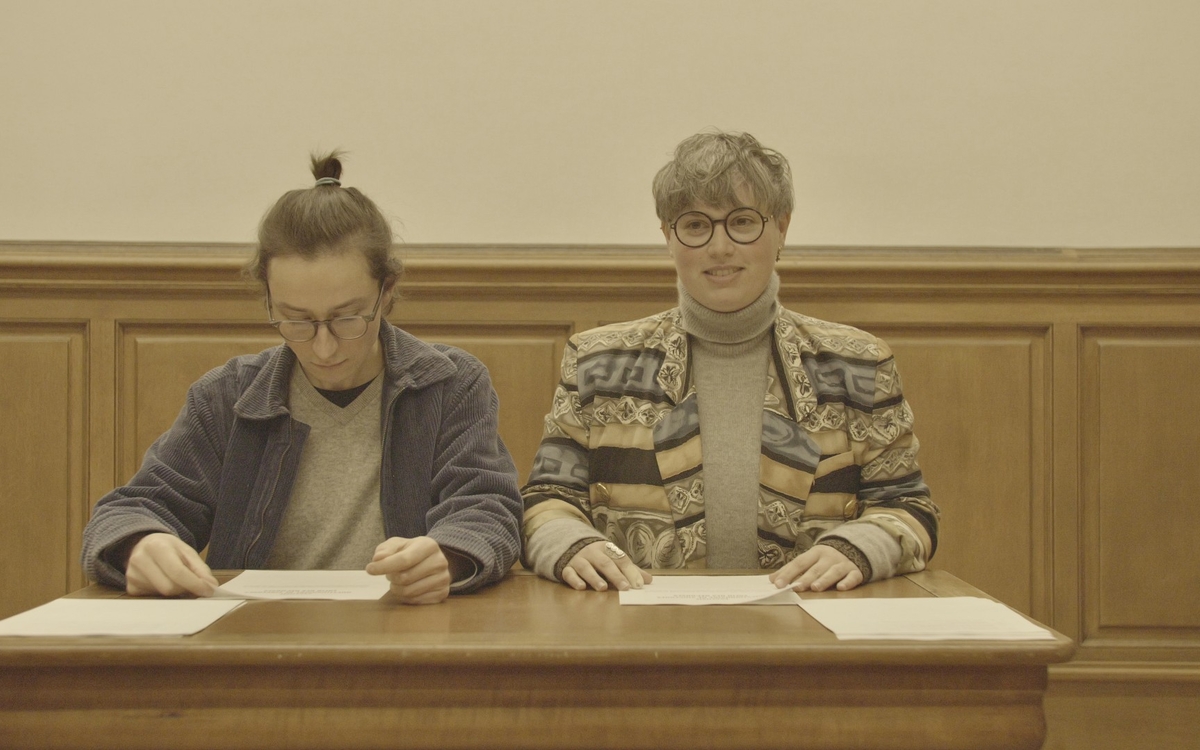 Deux artistes de la Compagnie les BdThé sont assises à un bureau face au public, l'une consulte des documents, l'autre regarde le public