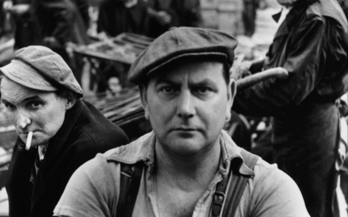 Photographie en noir et blanc représentant un homme au visage dur, les bras croisés