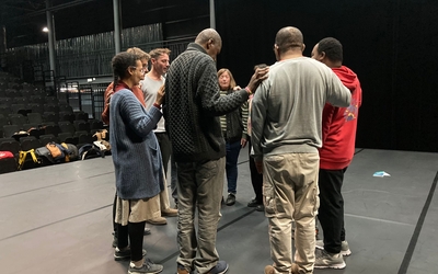 On y voit 8 personnes, adultes, en cercle sur un plateau de théâtre. Chacune a sa main droite posée sur l'épaule gauche de son ou sa voisine de droite. Elles ferment les yeux, ont l'air très concentré, dans un état méditatif. 