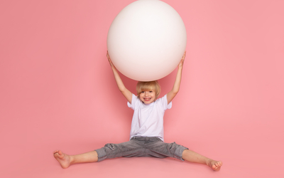 L'image représente un enfant assis portant un ballon de yoga gris. l'arrière plan est de couleur rose