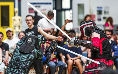 Deux combattants en tenue de protection affrontent leurs sabres lors d'un tournoi de sabre laser sportif en plein air, devant un public attentif.