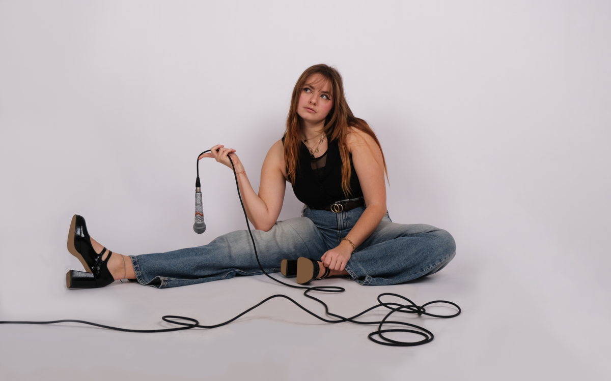 Chanteuse églantine assise par-terre avec un micro à la main, le regard évasif, sur fond blanc