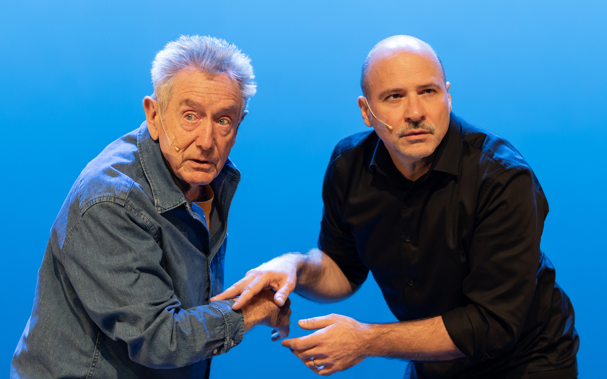 Les deux artistes sont devant un fond bleu, et se touchent les mains