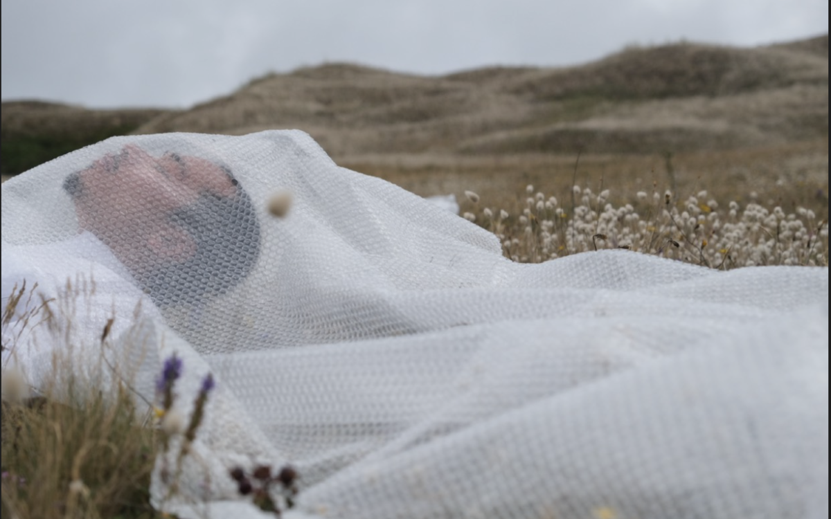 Un homme couché dans un champ les yeux fermés est recouvert d'un  tissu blanc transparent