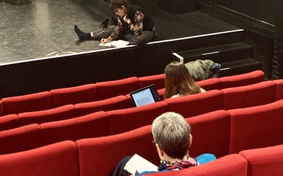 Trois personnes assises dans une salle de théâtre lisent chacunes de leur côté