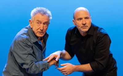 Les deux artistes sont devant un fond bleu, et se touchent les mains