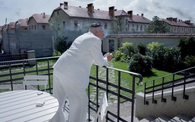 Un homme vêtu d'un costume blanc est penché sur la balustrade d'un porche, contemplant son jardin idyllique tout en fumant. À l'arrière-plan, on aperçoit les bâtiments d'un camp de concentration.