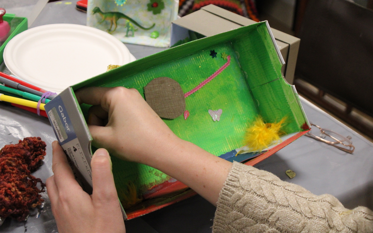 Photographie de mains d'enfants en train de confectionner un diorama avec de la peinture, des figurines, des paillettes, des plumes etc. dans une boîte à chaussures.