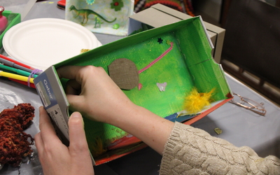 Photographie de mains d'enfants en train de confectionner un diorama avec de la peinture, des figurines, des paillettes, des plumes etc. dans une boîte à chaussures.