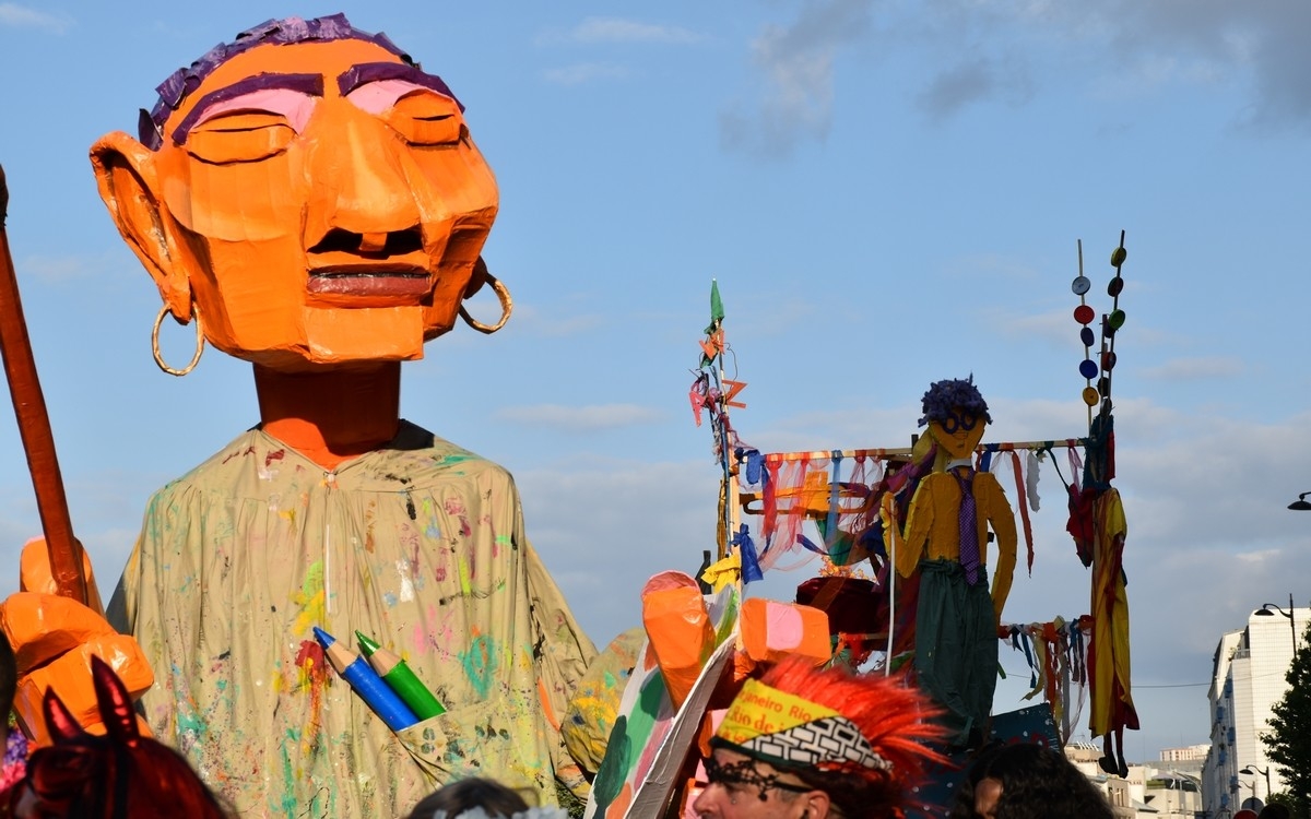 La photo représente un géant très coloré, avec un visage orangé, une blouse de travail avec des taches de peintures et 2 crayons de couleurs , un vert et un bleu sortent de sa poche située au niveau du coeur. Il tient une palette de couleurs dans sa main gauche et un énorme pinceau dans sa main droite. Derrière lui,  dans l'ombre, se trouve un autre personnage avec une chemise jaune et pantalon sombre, des cheveux noirs accroché à un char, où sont accrochés des bandes de tissus de toutes les couleurs