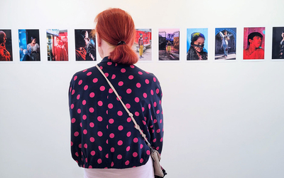 Une personne aux cheveux roux, vue de dos, observe une rangée de photographies colorées de l'artiste Thierry Chantegret accrochées sur un mur blanc dans une galerie. Elle porte une veste sombre à pois roses et un sac en bandoulière.