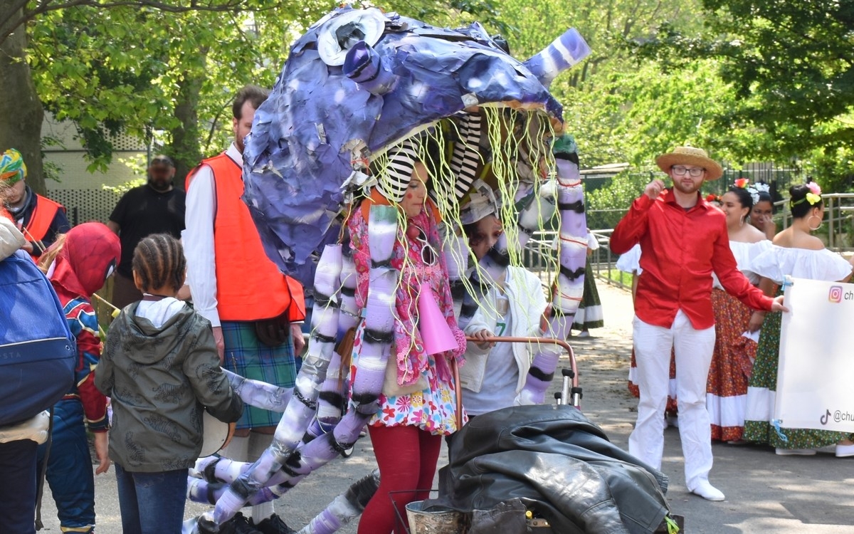 des adultes et des enfants en tenues festives sont autour d'une carnaval, de couleur bleutée. Elle représente une grosse pieuvre, portée par 2 personnes. 2 autres personnes sont en gilet orange pour encadrer leur sécurité.
