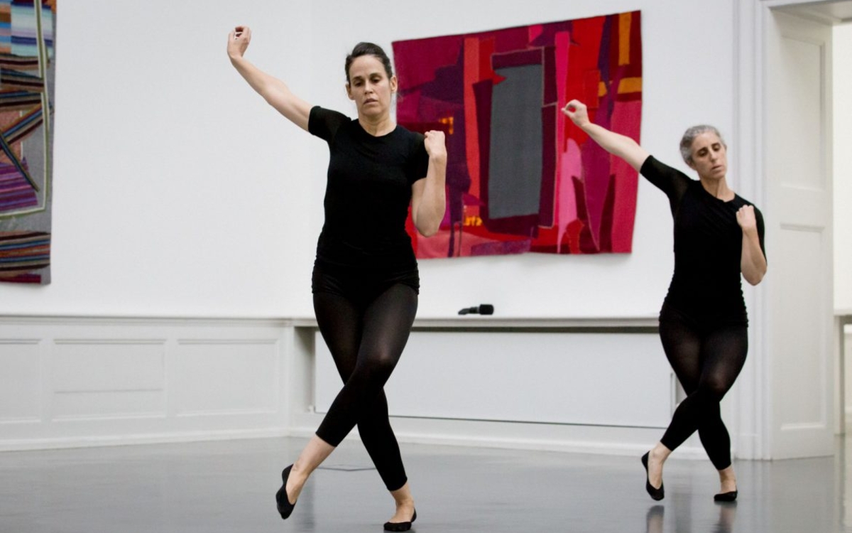 Deux danseuses effectuant des mouvements synchronisés, jambes croisées, le bras droit levé sur le côté, l'autre replié, le poing sur l'épaule, dans une pièce avec des œuvres textiles au mur.
