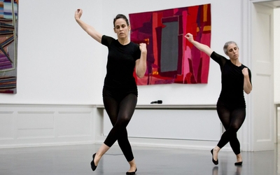 Deux danseuses effectuant des mouvements synchronisés, jambes croisées, le bras droit levé sur le côté, l'autre replié, le poing sur l'épaule, dans une pièce avec des œuvres textiles au mur.
