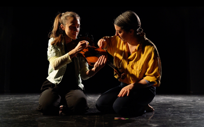 Sur cette image on voit deux femmes qui jouent du violon alto ensemble. l'une porte un chemisier jaune moutarde et l'autre un gilet blanc. Elles sont toute les deux assises sur les genoux et sont devant un fond noir.