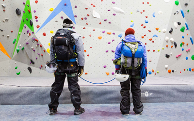 Deux interprètes sont face à un mur d'escalade avec des prises colorées. Ils sont vêtus d'une combinaison de ski et portent lunettes de soleil et bonnets. Ils portent également de gros sacs à dos et sont reliés entre eux par des cordes. 