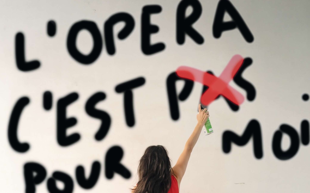 Une femme en robe rouge de dos raye le mot PAS à la bombe rouge au milieu de la phrase taguée au mur "L'Opéra c'est pas pour moi".