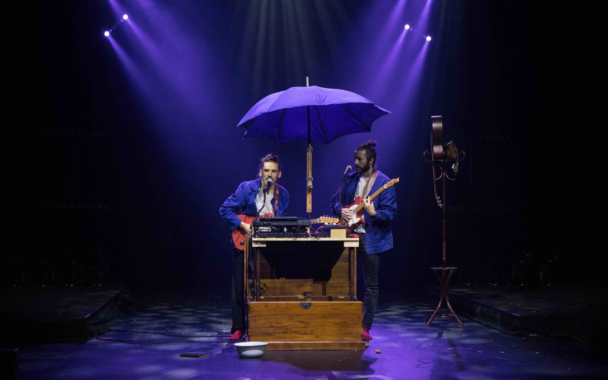 deux musiciens sur scène avec des guitares à la main, devant un instrument de leur fabrication fait entre autre de clavier, le tout sous un parapluie violet. 