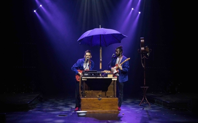 deux musiciens sur scène avec des guitares à la main, devant un instrument de leur fabrication fait entre autre de clavier, le tout sous un parapluie violet. 