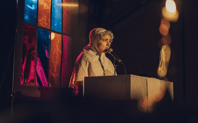 Photo d'une pièce de théâtre représentant une femme parlant dans un micro dans un décor qui semble celui d'une église