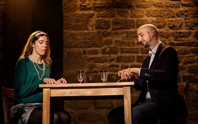 Deux adultes jouent une scène de théâtre face à face, devant un mur de briques.