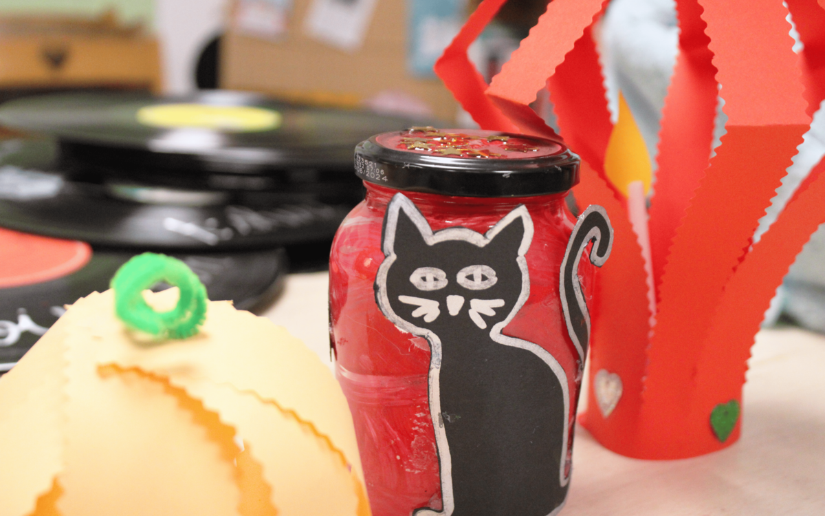 Lanterne peinte en rouge avec un dessin de chat noir fait à la main à la peinture. A côté se trouve des papiers colorés rouge et orange. 