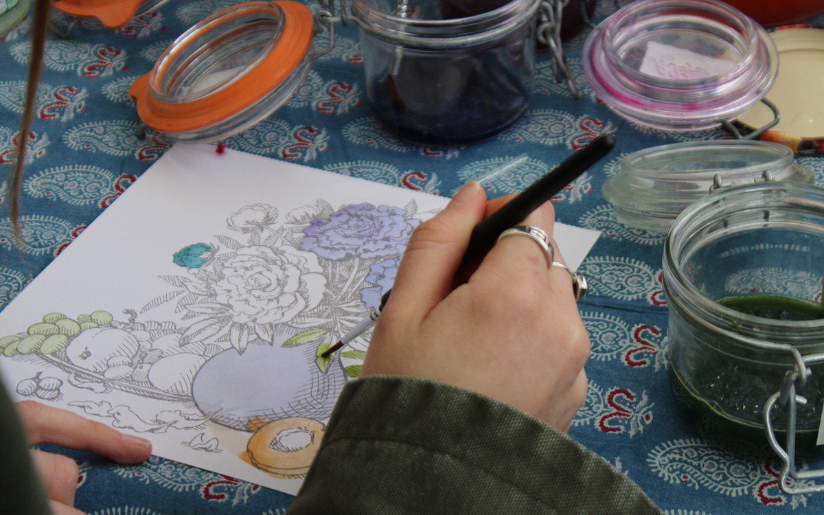 Photographie d'une main tenant un pinceau, en train de peindre un dessin de bouquet de fleurs avec de l'encre qui a été fabriquée à partir de végétaux. 